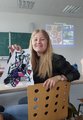 Eine lächelnde Schülerin sitzt auf einem Stuhl im Klassenzimmer und hält ein buntes Bild in die Kamera.