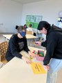 Eine Schülerin sitzt mit einer Augenbinde an einem Tisch. Eine andere Schülerin steht an der anderen Seite des Tisches und reicht der Mitschülerin Gegenstände an, die sie erfühlen soll.