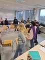 Die Klasse steht außen um einen Stuhlkreis herum. Sie halten die Stühle nach hinten gekippt und versuchen, den Stuhlkreis von Stuhl zu Stuhl zu umrunden.