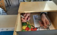 Das Bild zeigt eine Holzkiste, die vorne komplett und oben halb offen ist. Von Vorne hält ein Schüler seine Hände hinein und versucht, mit Messer und Gabel, Plastikgemüse auf die Gabel zu schieben.