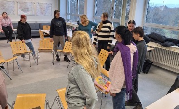 Die Klasse steht außen um einen Stuhlkreis herum. Sie halten die Stühle nach hinten gekippt und versuchen, den Stuhlkreis von Stuhl zu Stuhl zu umrunden.