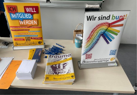 Auf dem Tisch stehen zwei bunte Aufsteller mit dem Logo des Berufsverband. Daneben lieben Stifte, Blöcke und Flyer des Verbands.