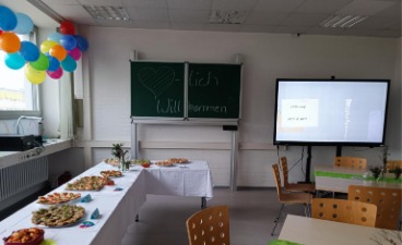 Das Foto zeigt ein Klassenzimmer am Tag der offenen Tür. Auf den Tischen stehen Teller mit Kuchen und Häppchen. Auf die Tafel ist "Herzlich Willkommen" geschrieben und das mobile Whiteboard zeigt eine Foto-Präsentation.