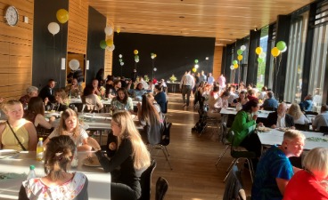 In einem großen, offenen Saal sitzen Gäste an Tischen zusammen und unterhalten sich. Die Tische sind mit Luftballons , Kerzen und Mooskugeln dekoriert.