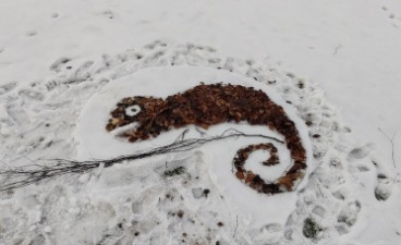 Das Bild zeigt eine Land-Art-Figur aus Stöcken und Blättern im Schnee. Sie stellt ein Chamäleon auf einem Ast dar.