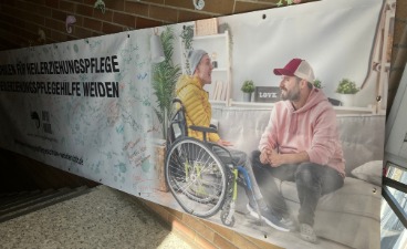 Das Bild zeigt ein langes Banner, das im Treppenhaus hängt und auf dem ein Rollstuhlfahrer, einen assistierende Fachkraft und der Name der Schule abgebildet sind.